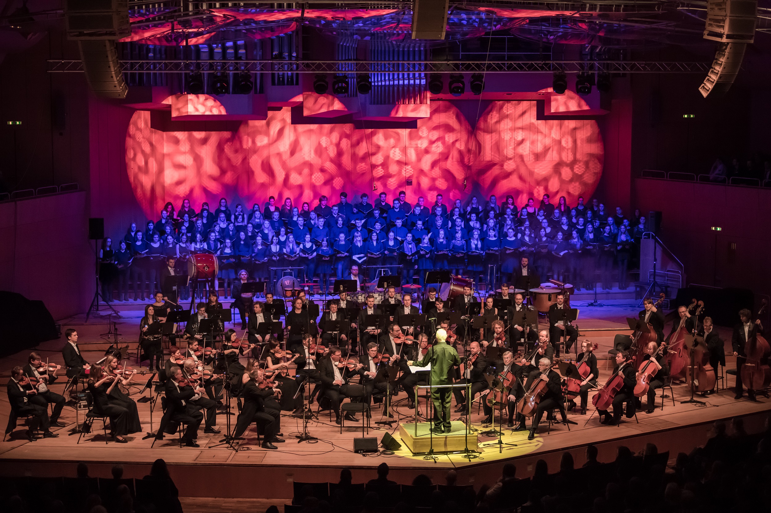 Das Czech Symphonie Orchestra und dahinter ein großer Chor sind auf einer Bühne zu sehen. Der Dirigent wird gelb, der Chor blau und der Hintergrund rot beleuchtet.