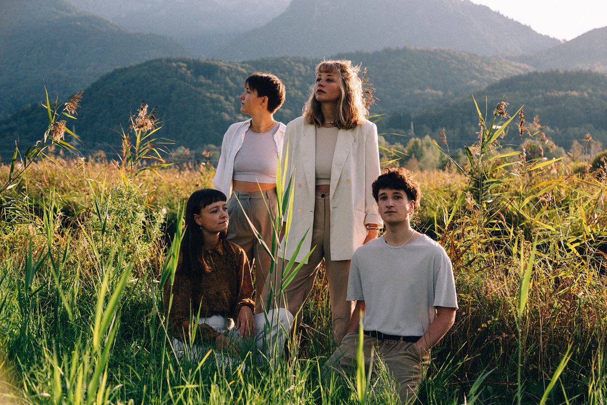 Three women and a man stand and squat in a grassy landscape. In the background you can see forested mountains.