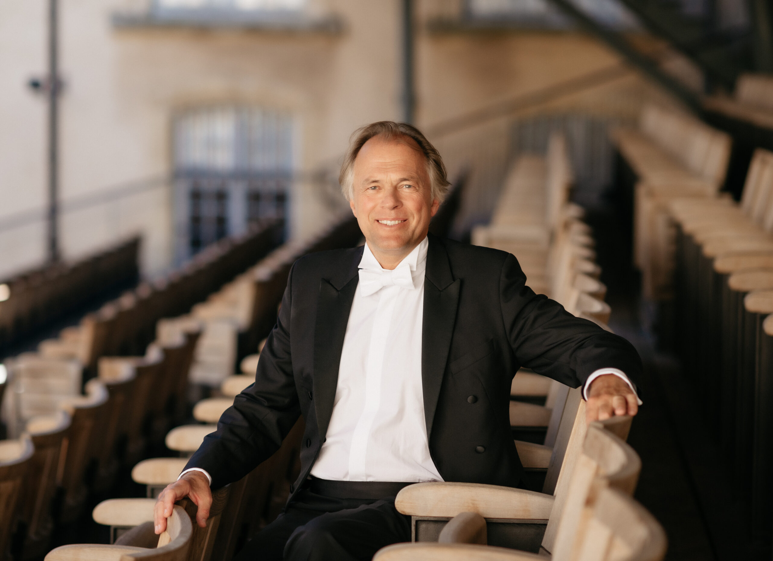 Conductor Thomas Hengelbrock sits in the auditorium and smiles at the camera
