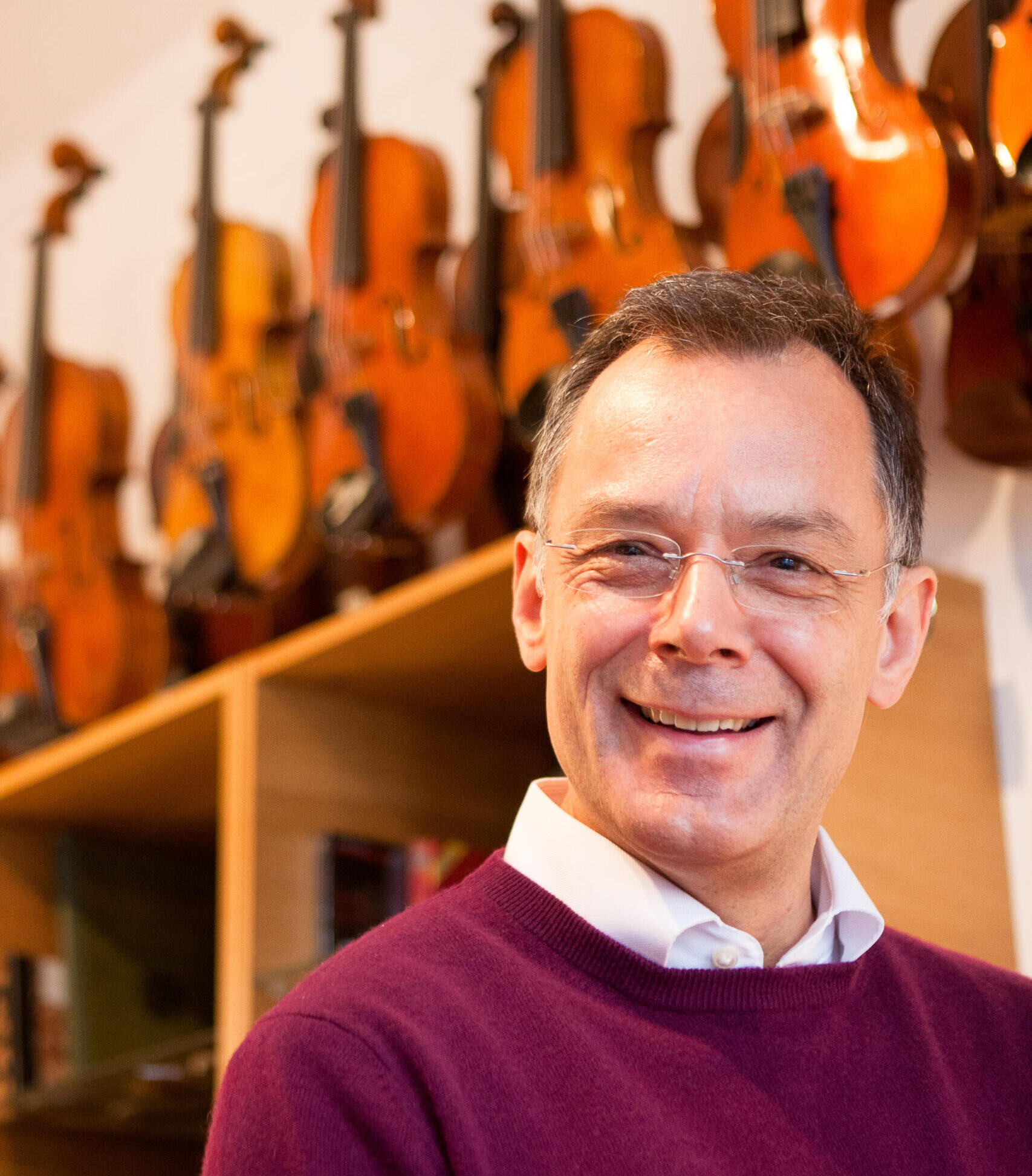 Portraitfoto des Geigenbauers Jörg Trautmann. Im Hintergrund sind diverse Geigen auf einem Regal zu sehen.