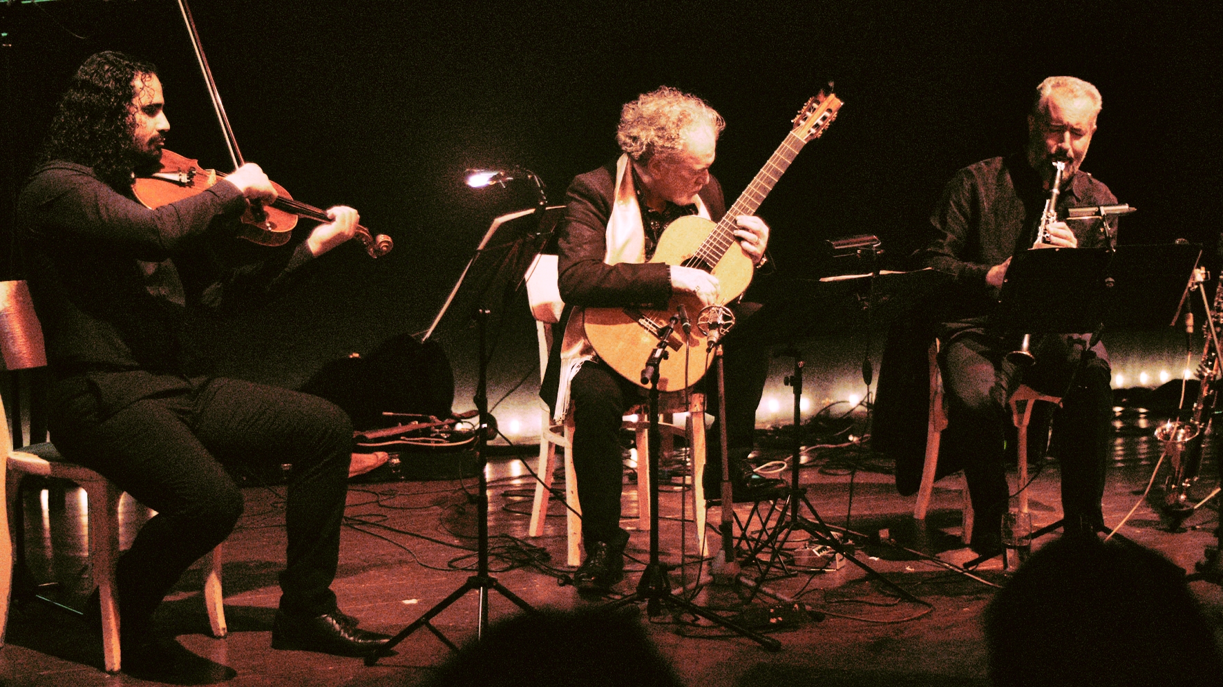 Drei Musiker sitzen auf einer Bühne und spielen ihre Instrumente, eine Bratsche, eine Klarinette und eine Gitarre.