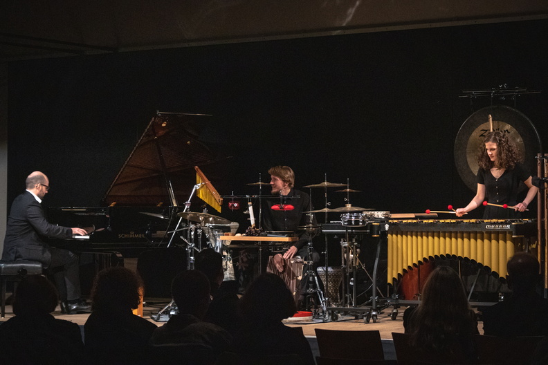 A stage with 3 musicians: A man on the grand piano, one on the drums and a woman on the xylophone