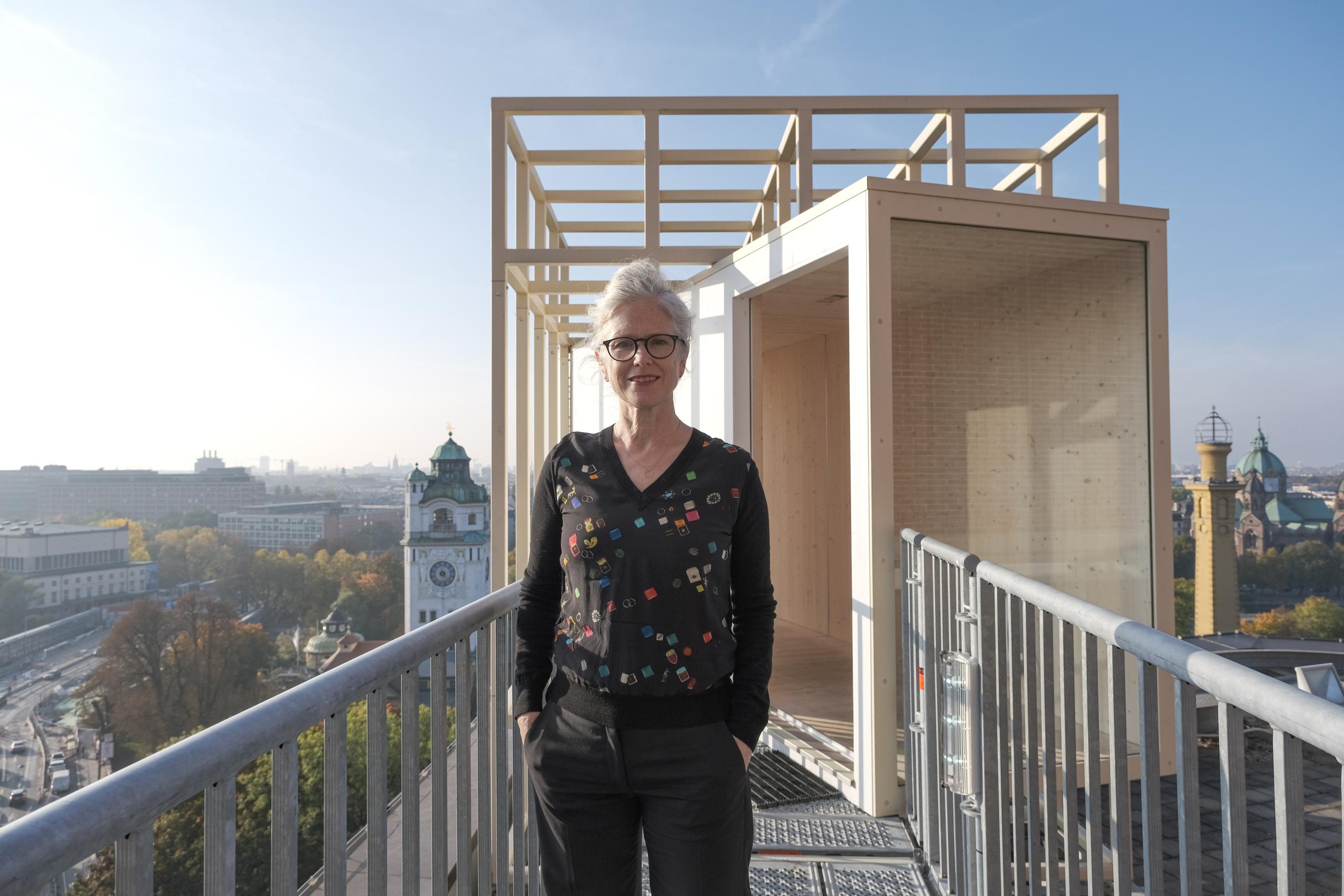 Eine Frau steht auf dem Gerüstzugang des Aussichtsraums über München und blickt in die Kamera.