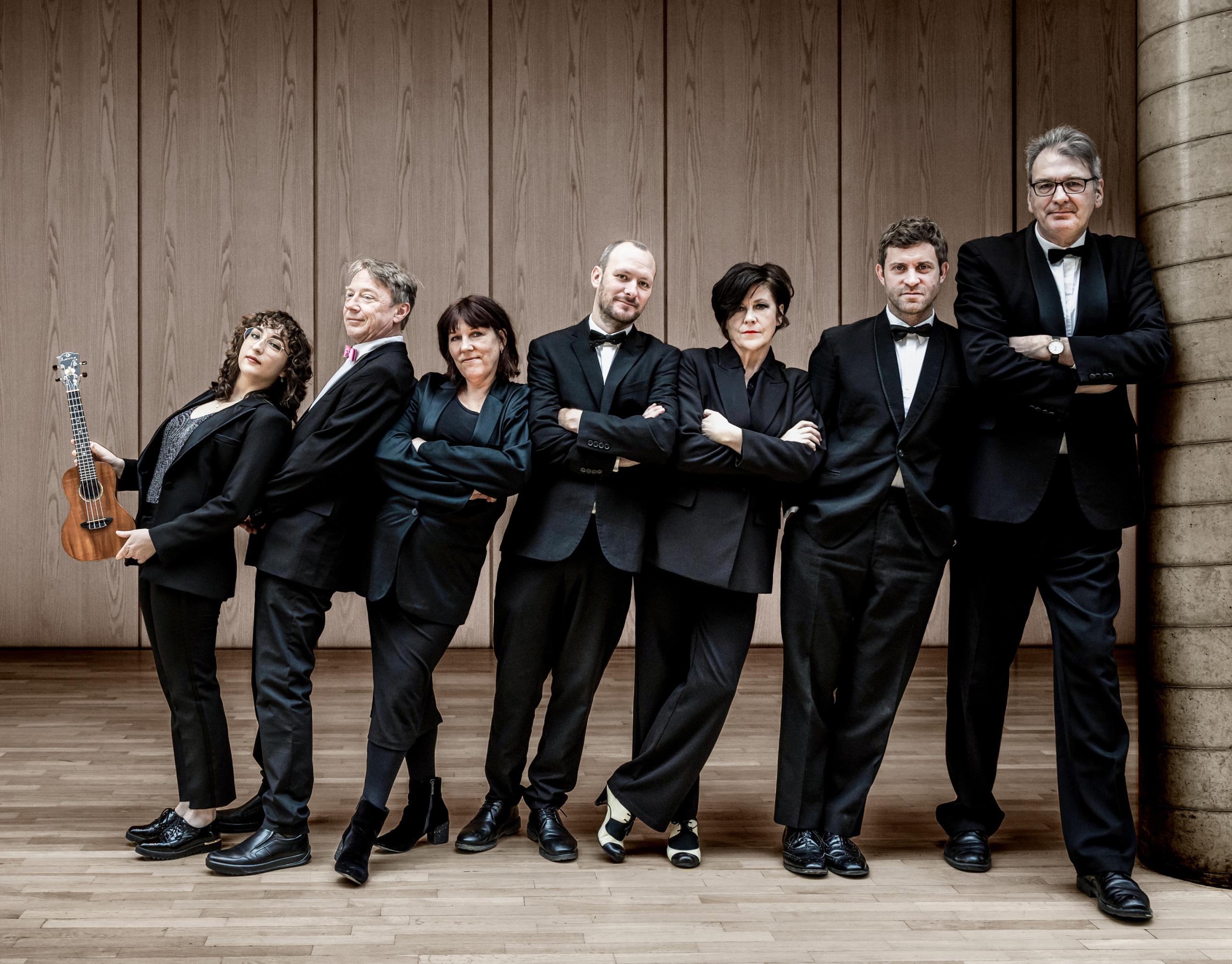 The members of the Ukulele Orchestra of Great Britain stand in black suits in front of a wooden wall and look into the camera.
