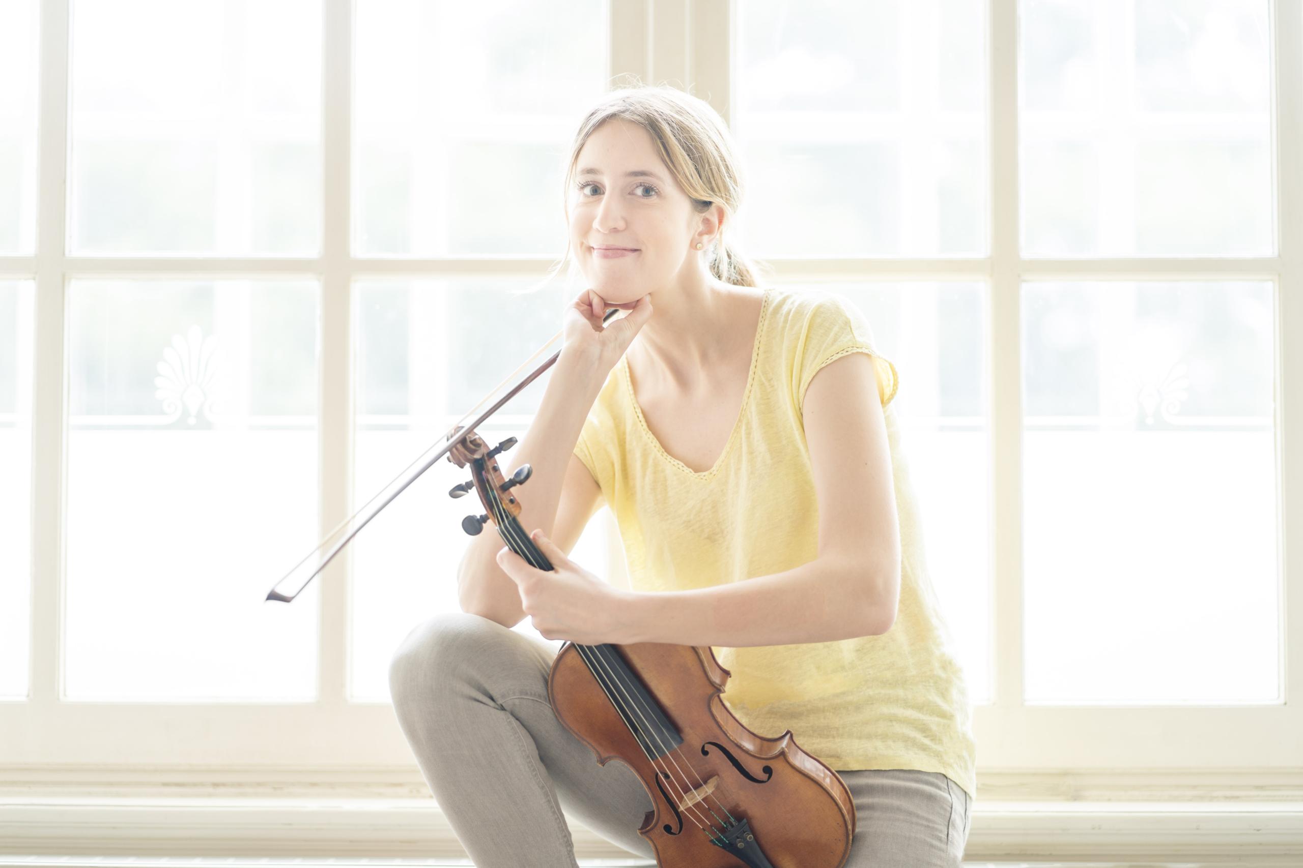 Die Geigerin Vilde Frang sitzt auf einer Fensterbank und stützt sich entspannt mit dem Ellenbogen auf dem Knie ab. Den Bogen und die Geige hält sie in jeweils einer Hand.