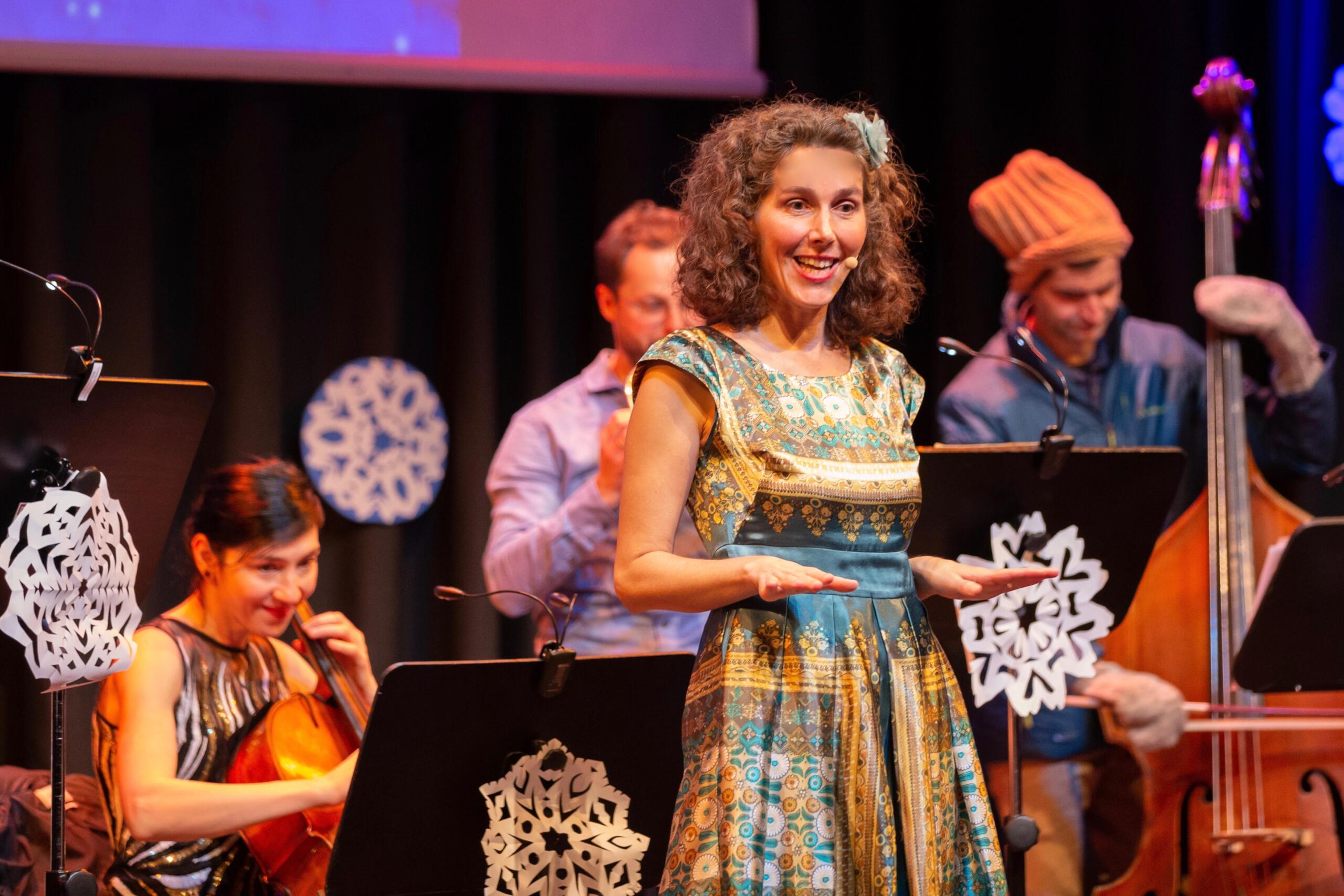 Eine Frau in einem gemusterten Kleid steht auf einer Bühne. Im Hintergrund sieht man Musiker und Musikerinnen mit ihren Instrumenten.