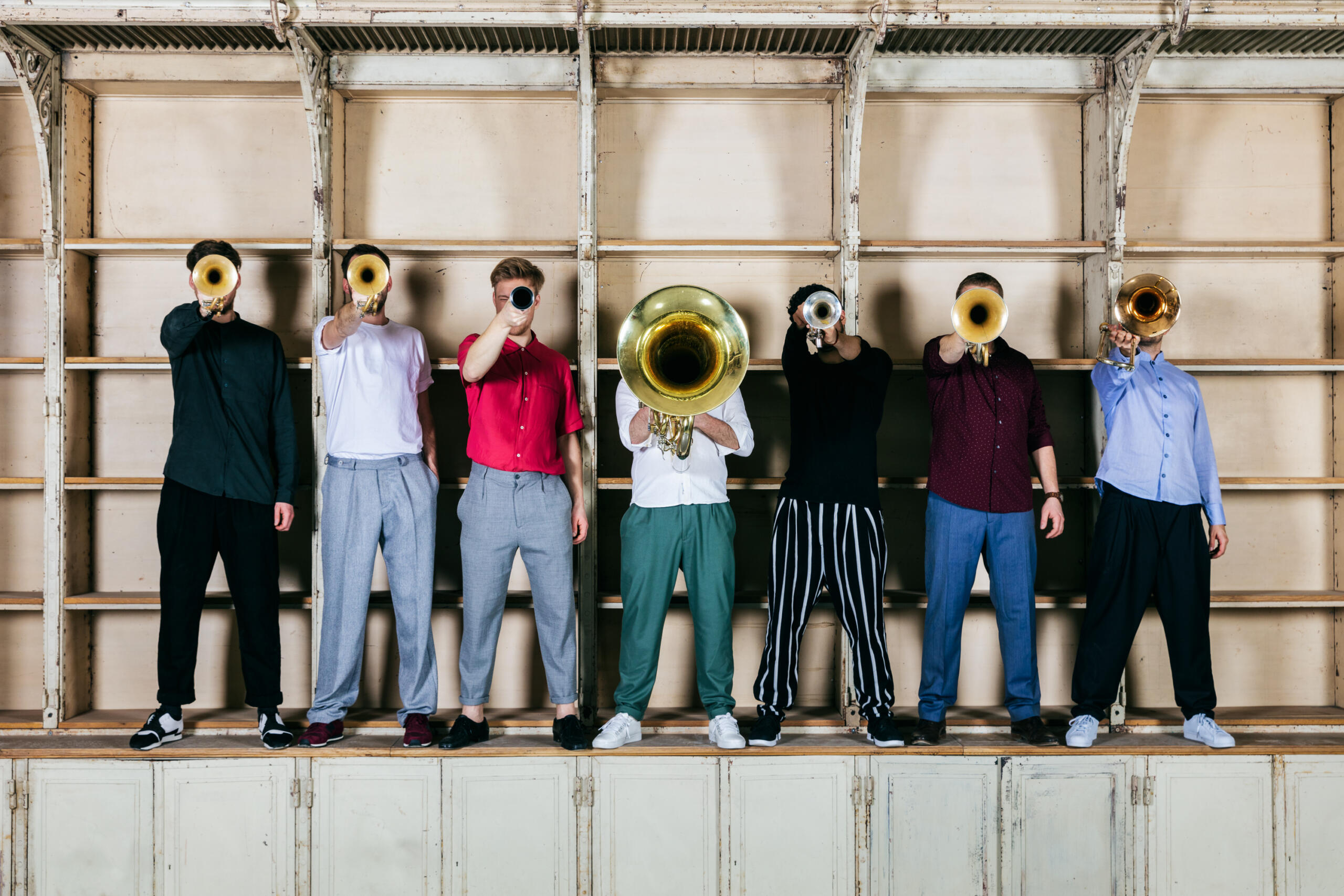 Sieben Musiker stehen in einer Reihe und halten ihre Blasinstrumente so, dass ihre Gesichter nicht zu sehen sind.