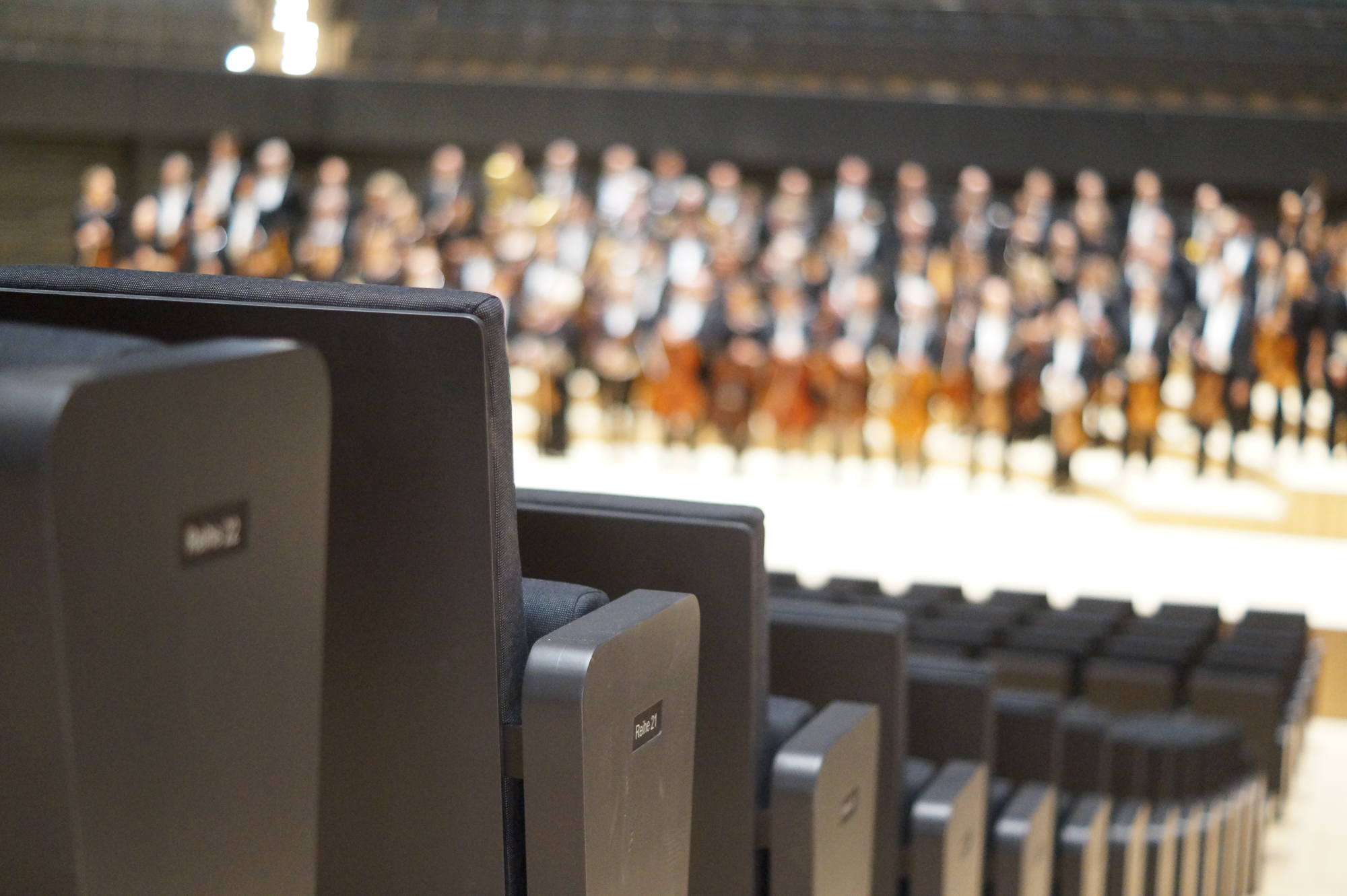 Leere Sitzreihen, man blickt auf die Bühne, hier sieht man unscharf die Münchner Philharmoniker