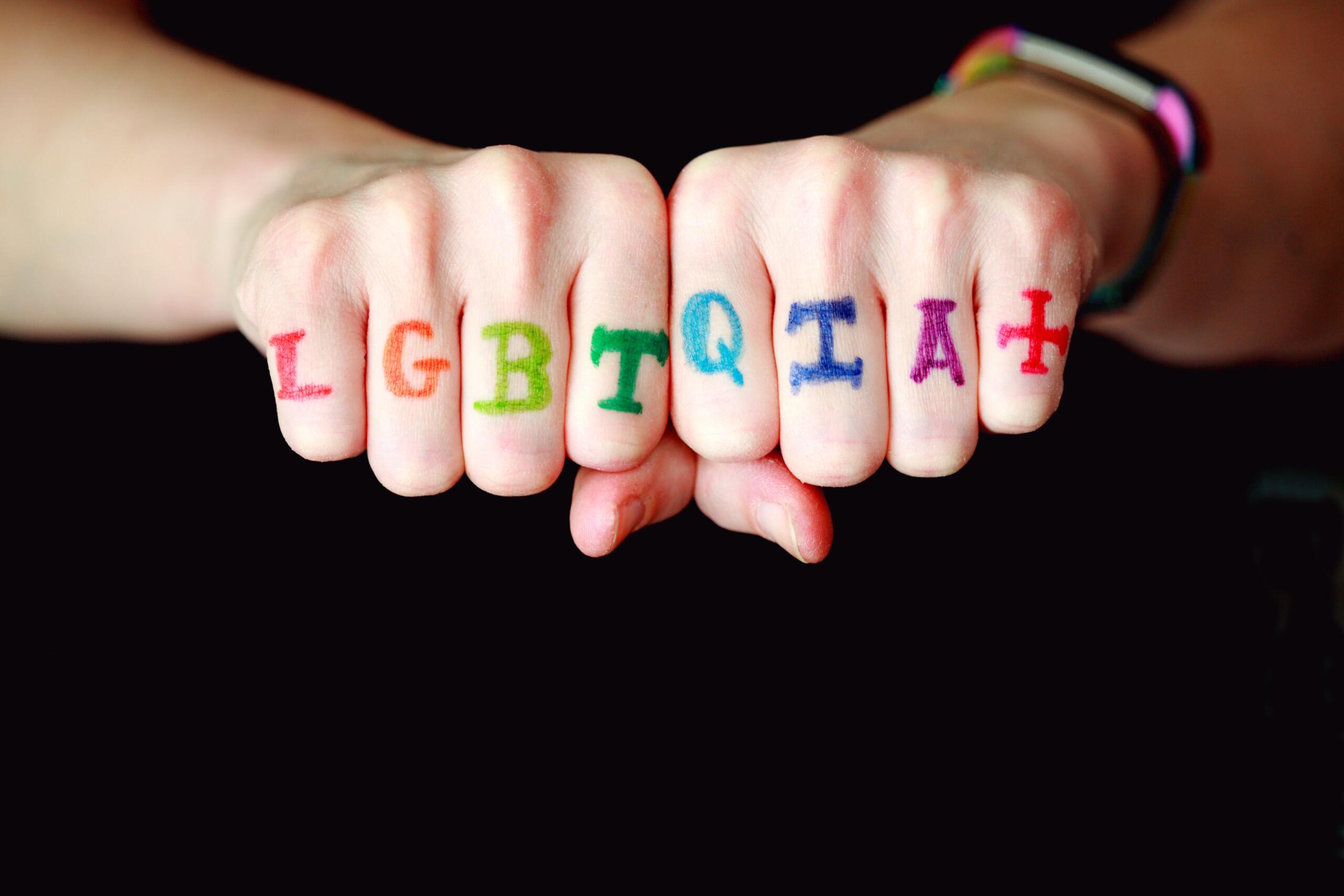 Zwei Fäuste nebeneinander, auf jeden Finger ist in unterschiedlichen Farben ein Buchstabe von LQBTQIA+ geschrieben. 