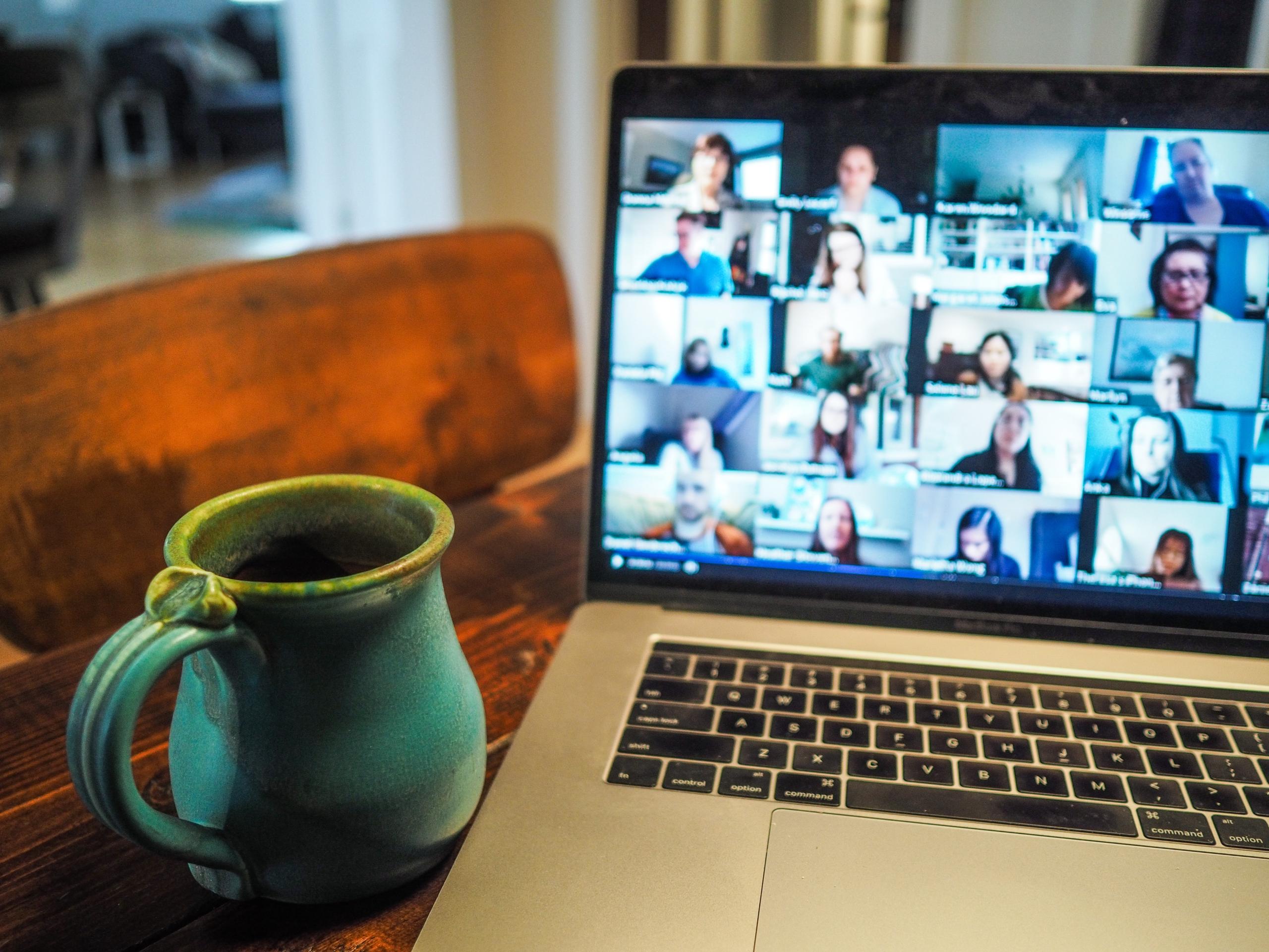 Laptop auf einem Holztisch, eine Tasse daneben, auf dem Bildschirm ein Videocall mit vielen Menschen