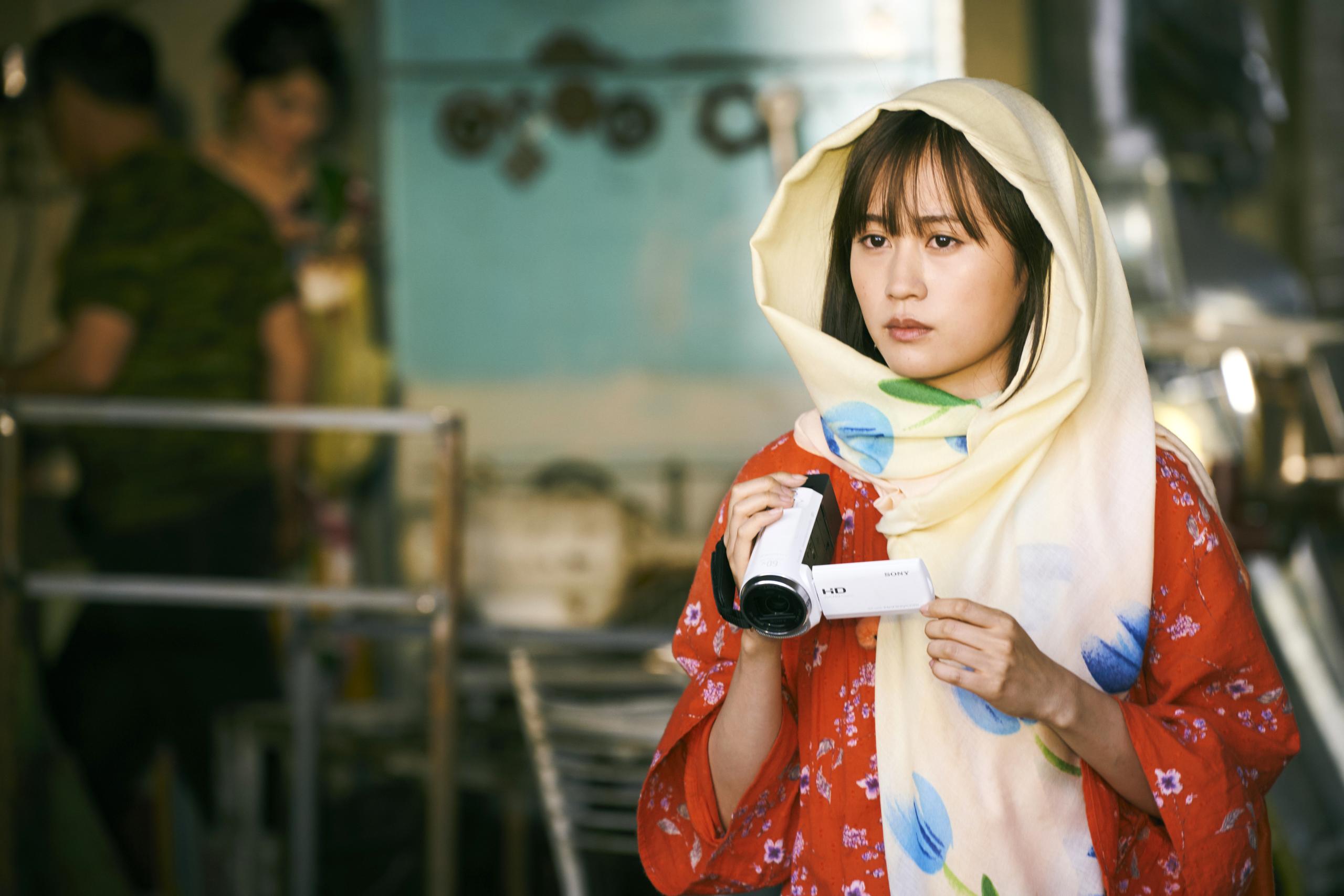 Szene auf einem Markt, eine Frau mit einem Schal um den Kopf und einer Handkamera schaut verträumt nach vorne