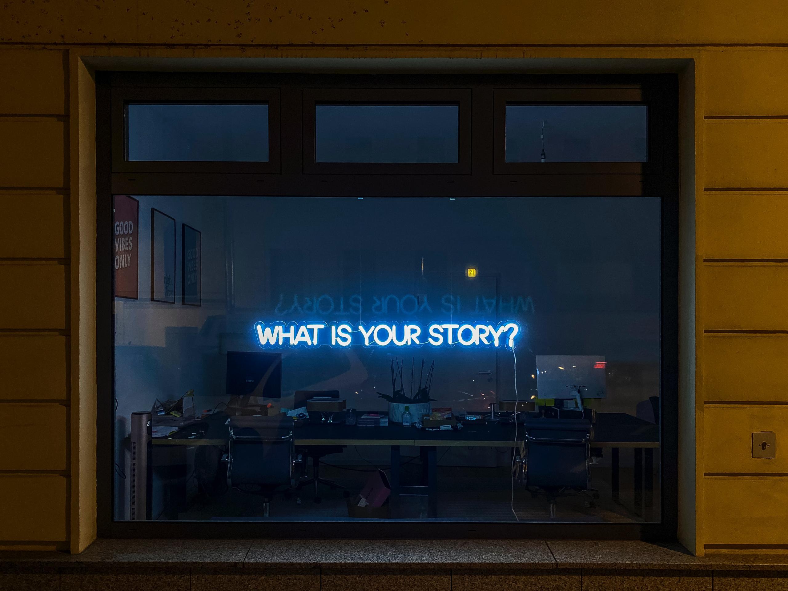 Schaufenster einer Agentur, man sieht Schreibtische im Hintergrund. Im Fenster steht in Neonschrift What is your story?