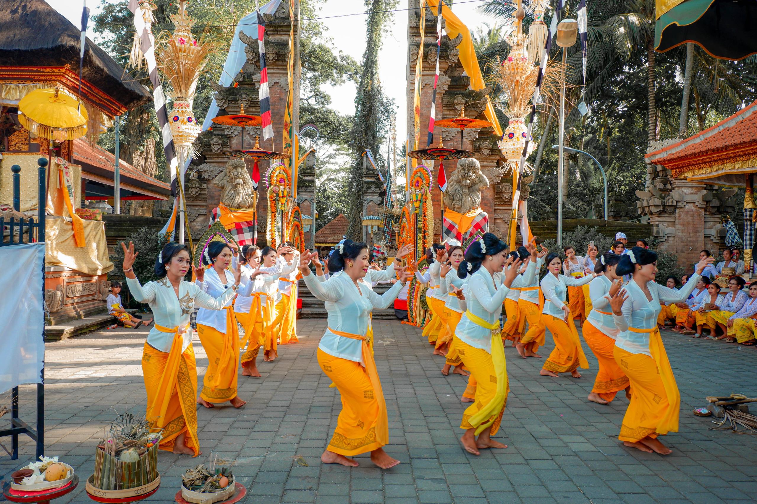 A Balinese temple, in front of it women dance in 4 rows. They wear white blouses and orange sarongs.