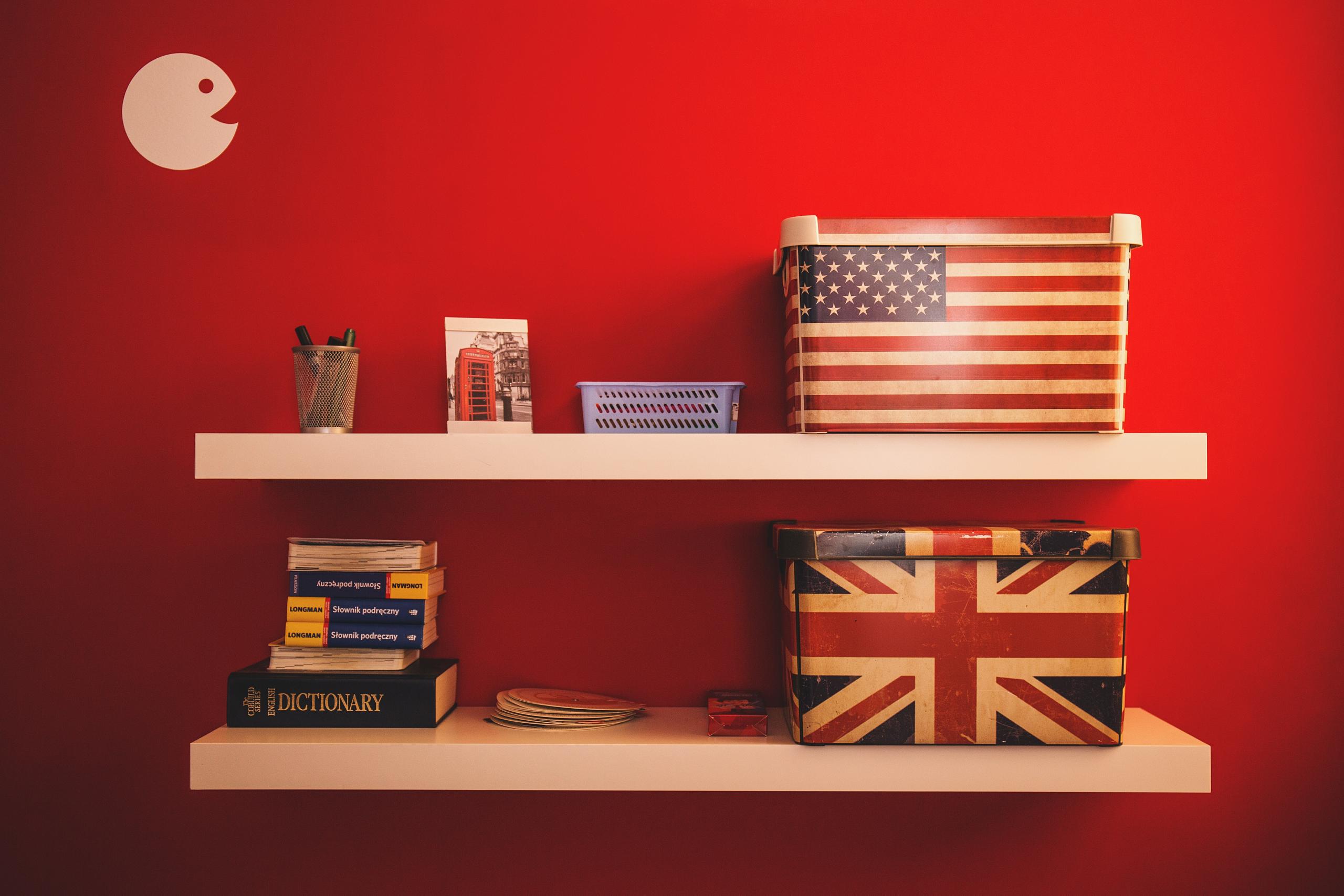Zwei Wandregale, auf denen Schachteln stehen, die eine ist mit der amerikanischen, die andere mit der englischen Fahne bedruckt. Daneben sind Wörterbücher gestapelt und Schreibsachen. Die Wand ist rot gestrichen.