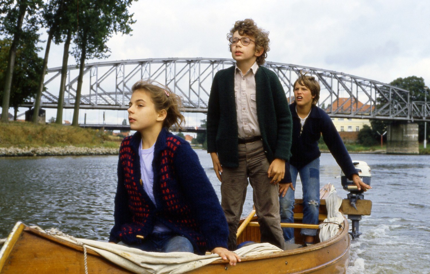 Zwei Jungen und ein Mädchen sitzen in einem Holzboot und fahren auf einem Fluss entlang.