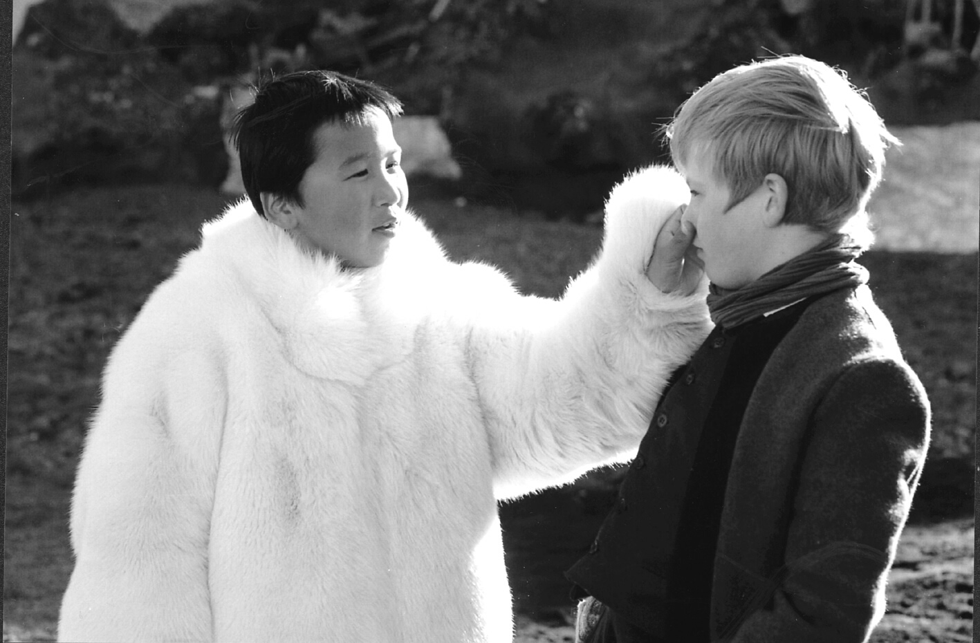 Eine schwarz-weiß Fotografie auf der in Junge in einem weißen Pelzmantel und ein Junge in einem dunklen schicken Mantel sich gegenüber stehen. Der Junge im Pelzmantel kneift den anderen Jungen in die Nase.