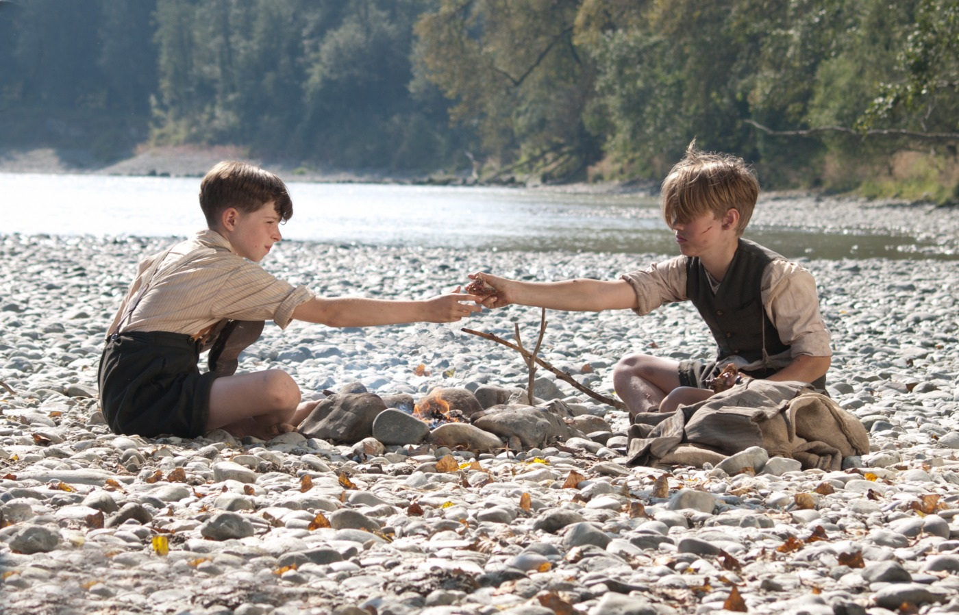 Zwei Jungen sitzen sich am Ufer eines Flusses gegenüber. Zwischen ihnen brennt ein Lagerfeuer.