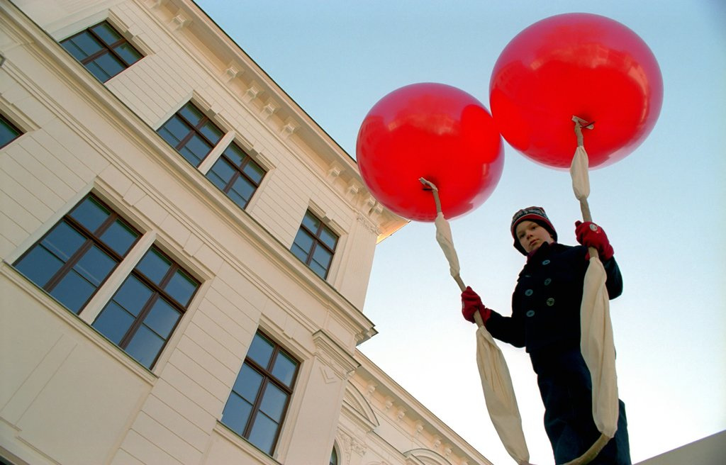 Ein Junge ist von unten zu sehen, wie er befestigt an zwei roten großen Heliumballons nach oben fliegt.