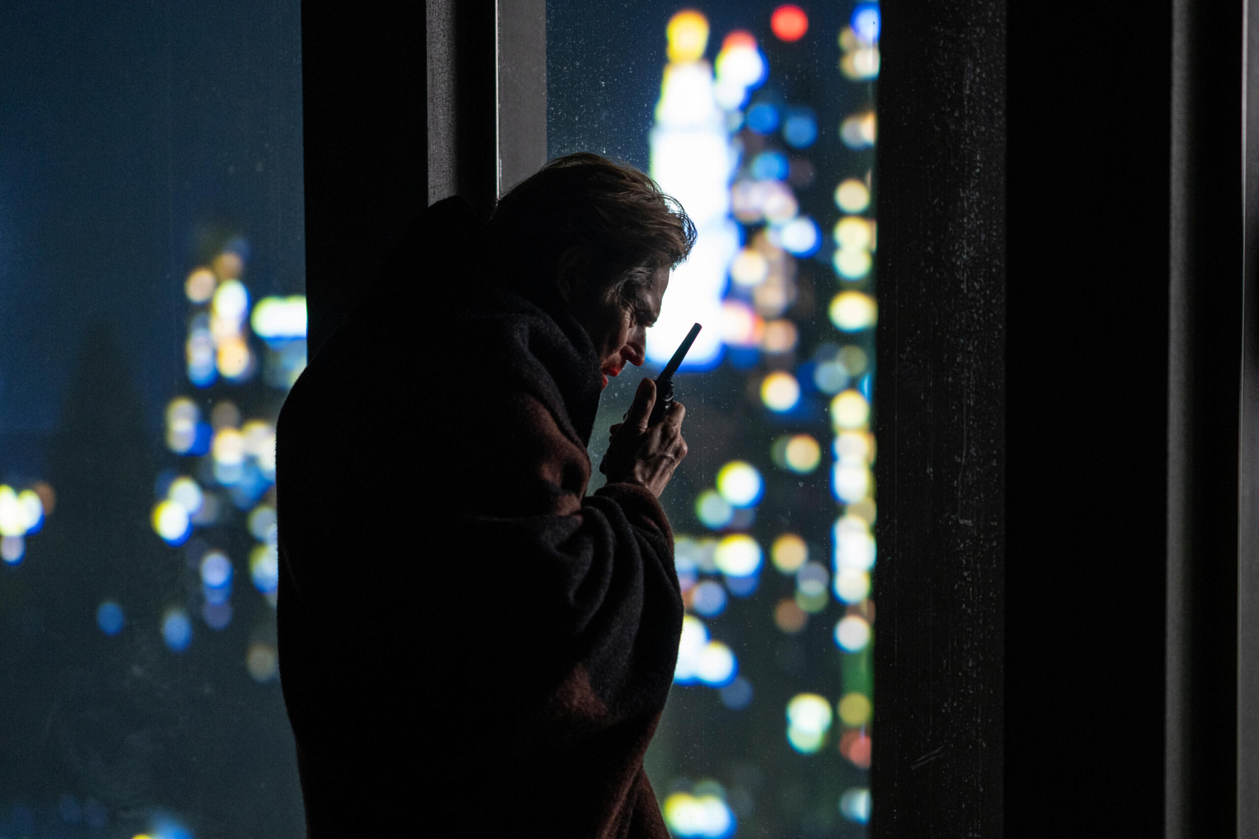 Ein Mann steht in eine Decke gehüllt vor einem Fenster, durch das schemenhaft die Skyline von New York zu erkennen ist.