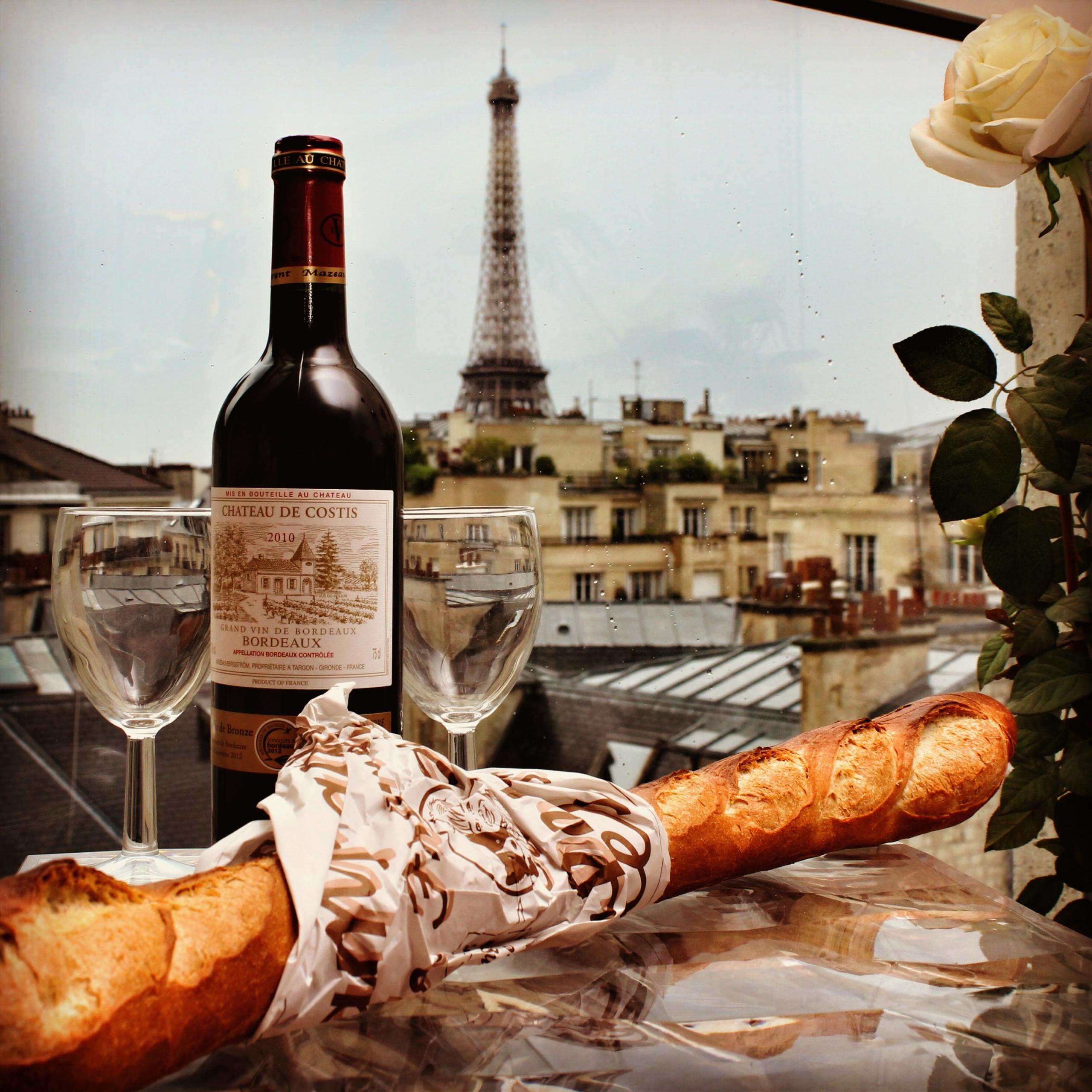 Blick über die Dächer von Paris zum Eiffelturm, vorne im Bild eine Flasche Bordeaux, zwei Weingläser und eine Baguette.