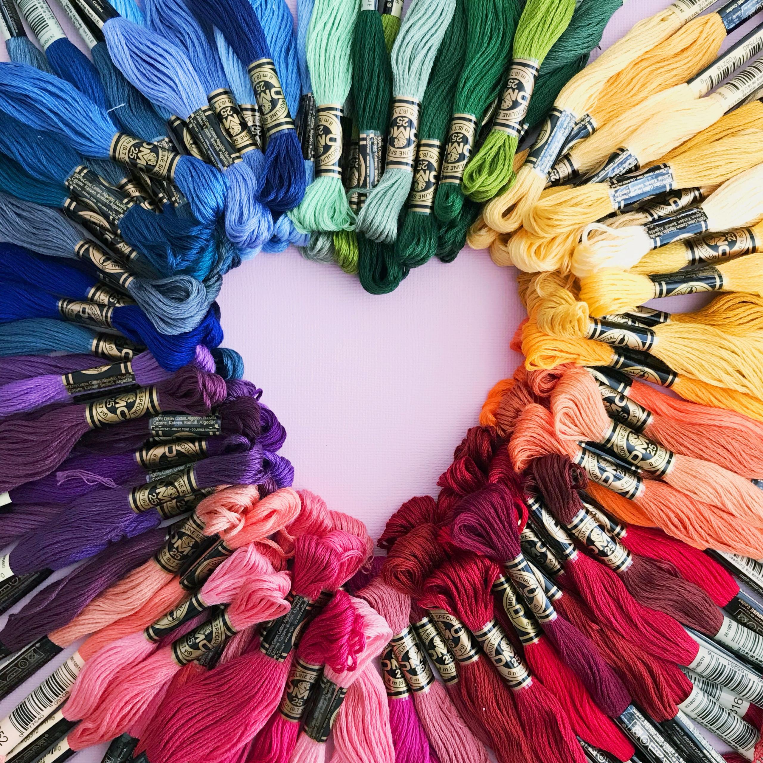 Viele Packungen Stickgarn sind farblich wie ein Regenbogen sortiert als Kreis gelegt. Die Mitte bildet ein Herz.