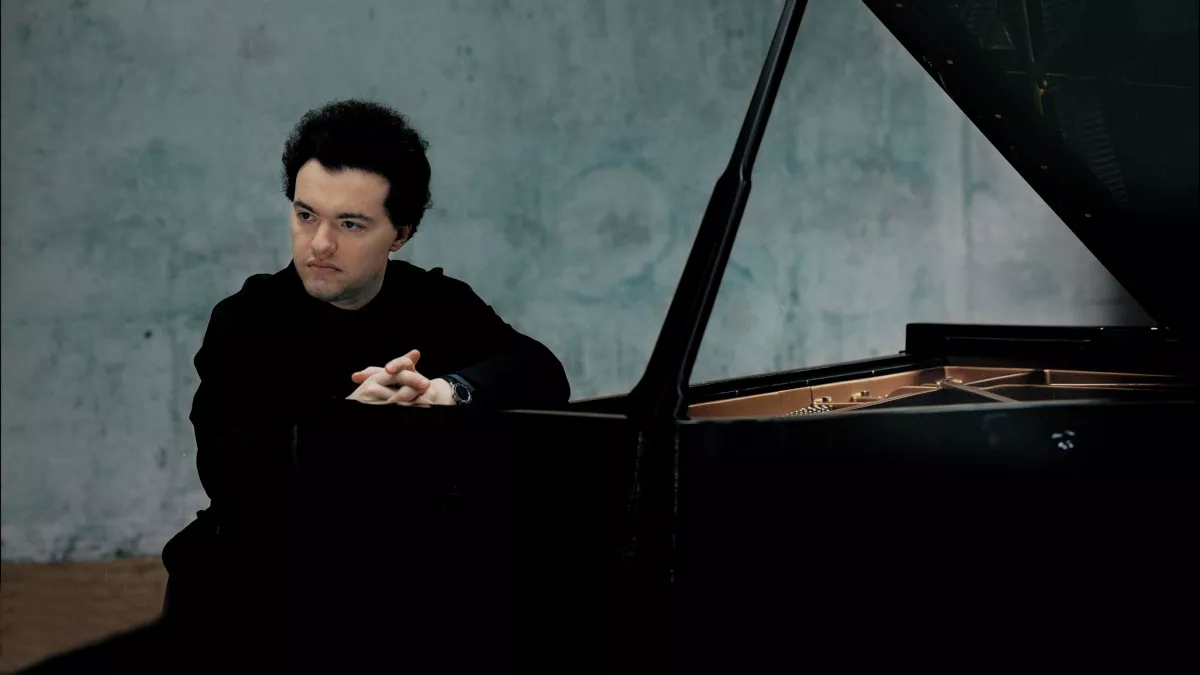 Portrait of the pianist Evgeny Kissin. He sits with folded hands behind a grand piano and looks pensively into the distance.