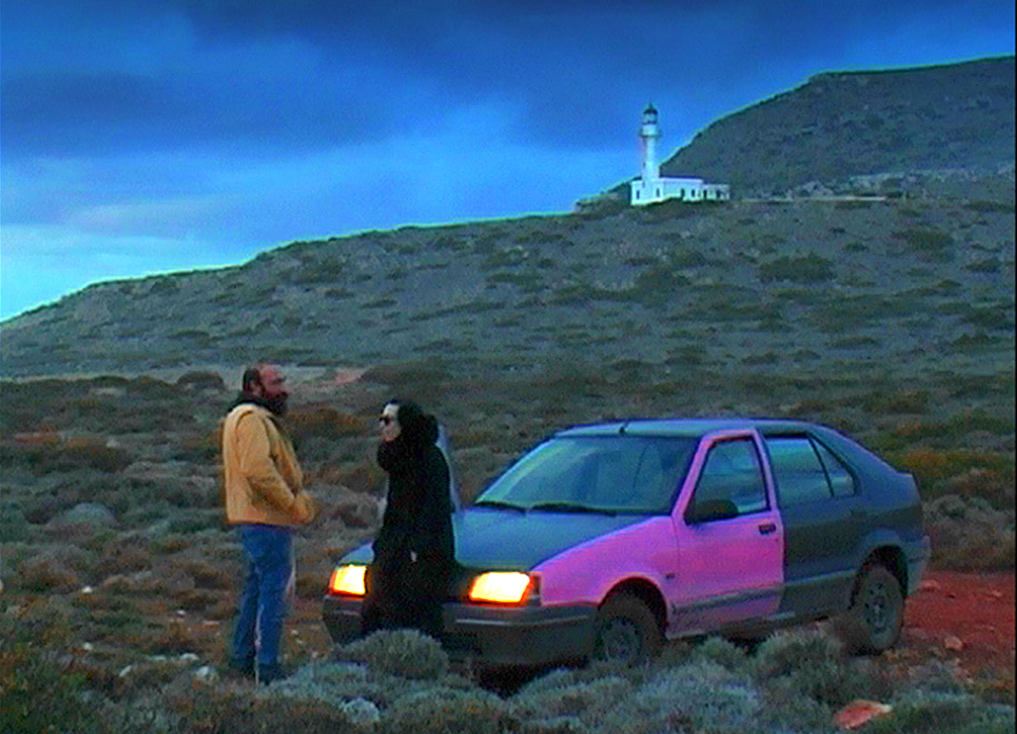 Eine Frau und ein Mann stehen vor einem Auto. Im Hintergrund sieht man eine Düne, einen Leuchtturm und dahinter den trüben Himmel.