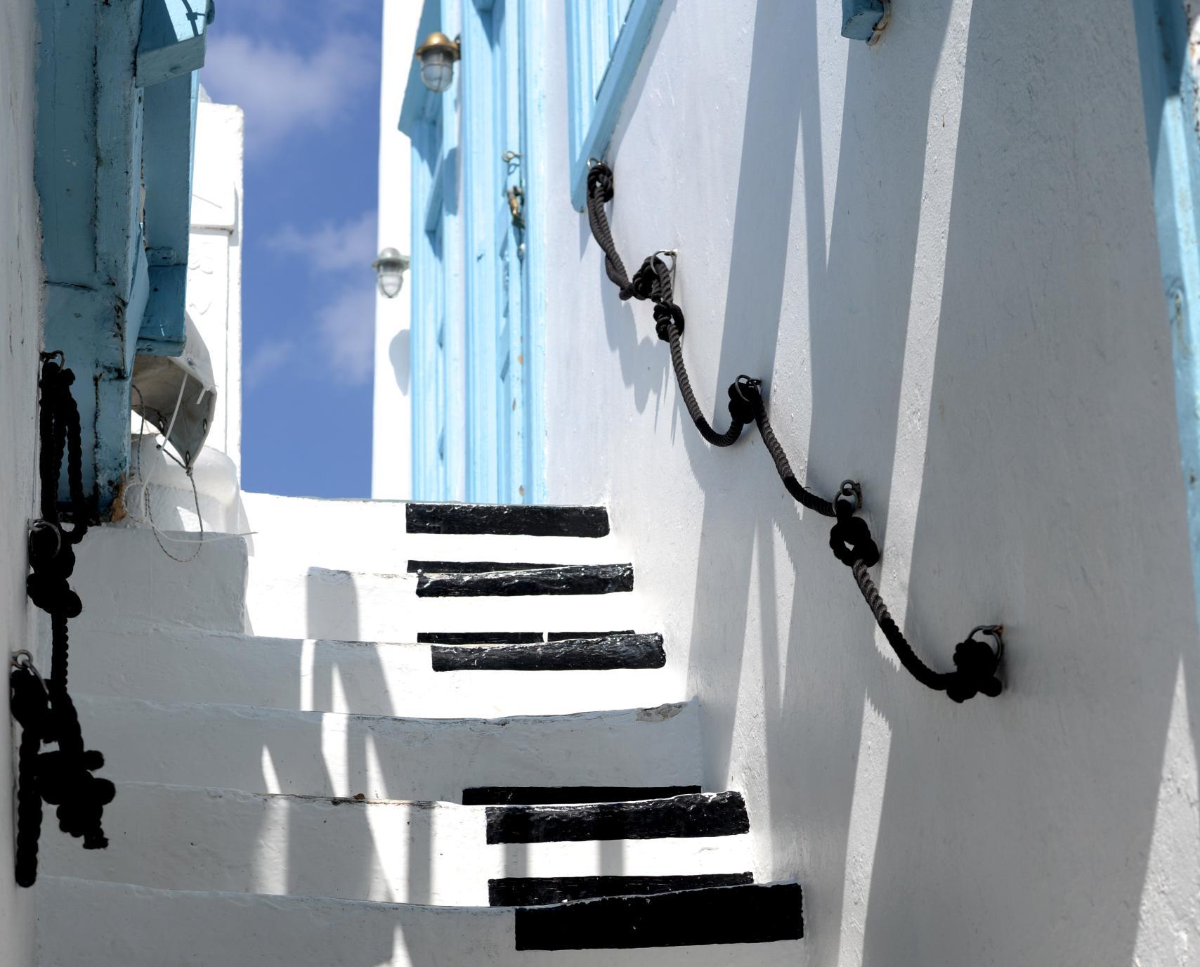 Greek alley, whitewashed walls, blue windows and doors. A staircase leads up, black piano keys are painted on the white steps.