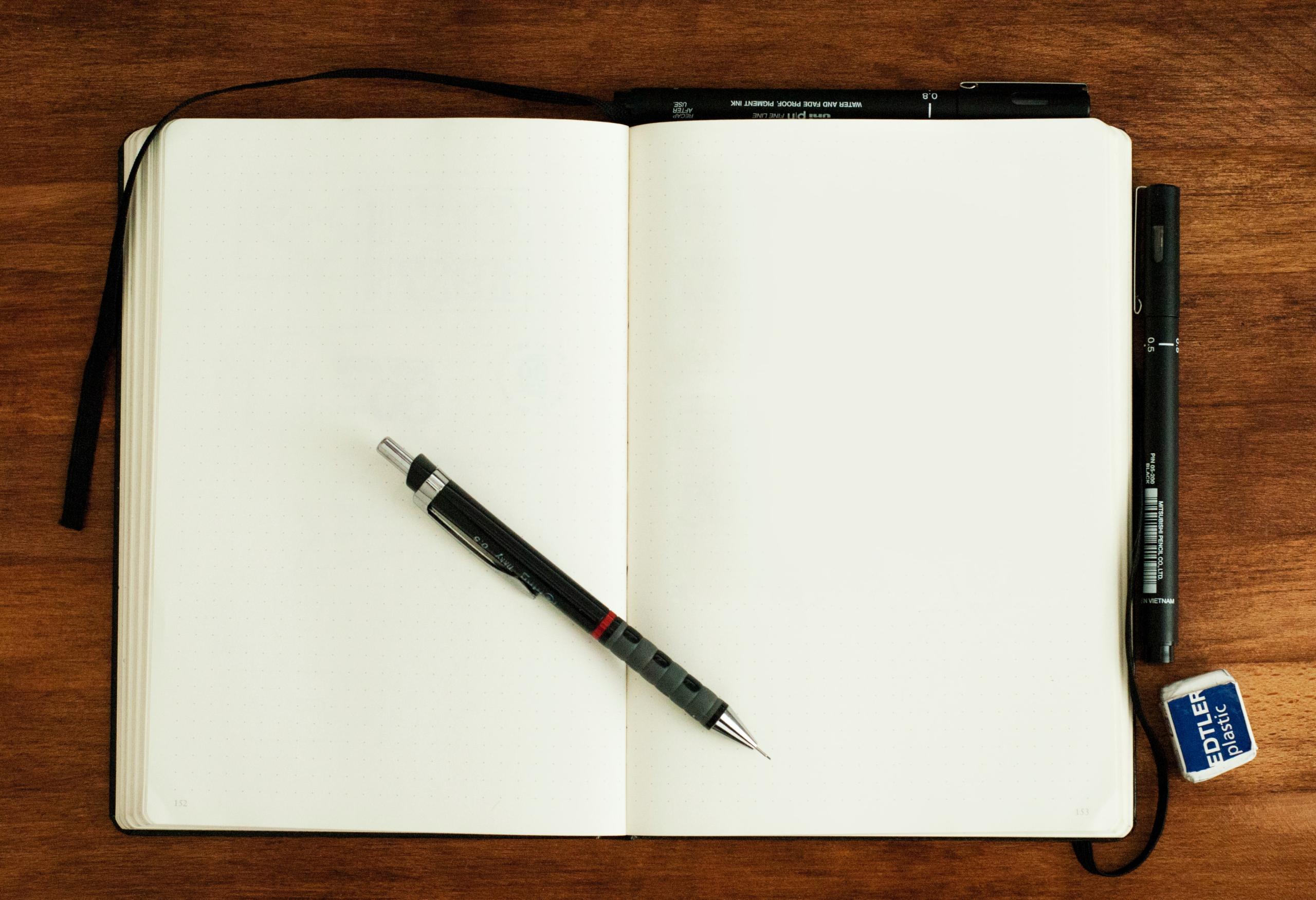 Ein leeres Skizzen- oder Notizbuch, darauf ein Bleistift, rundum weitere Stifte und Radiergummi