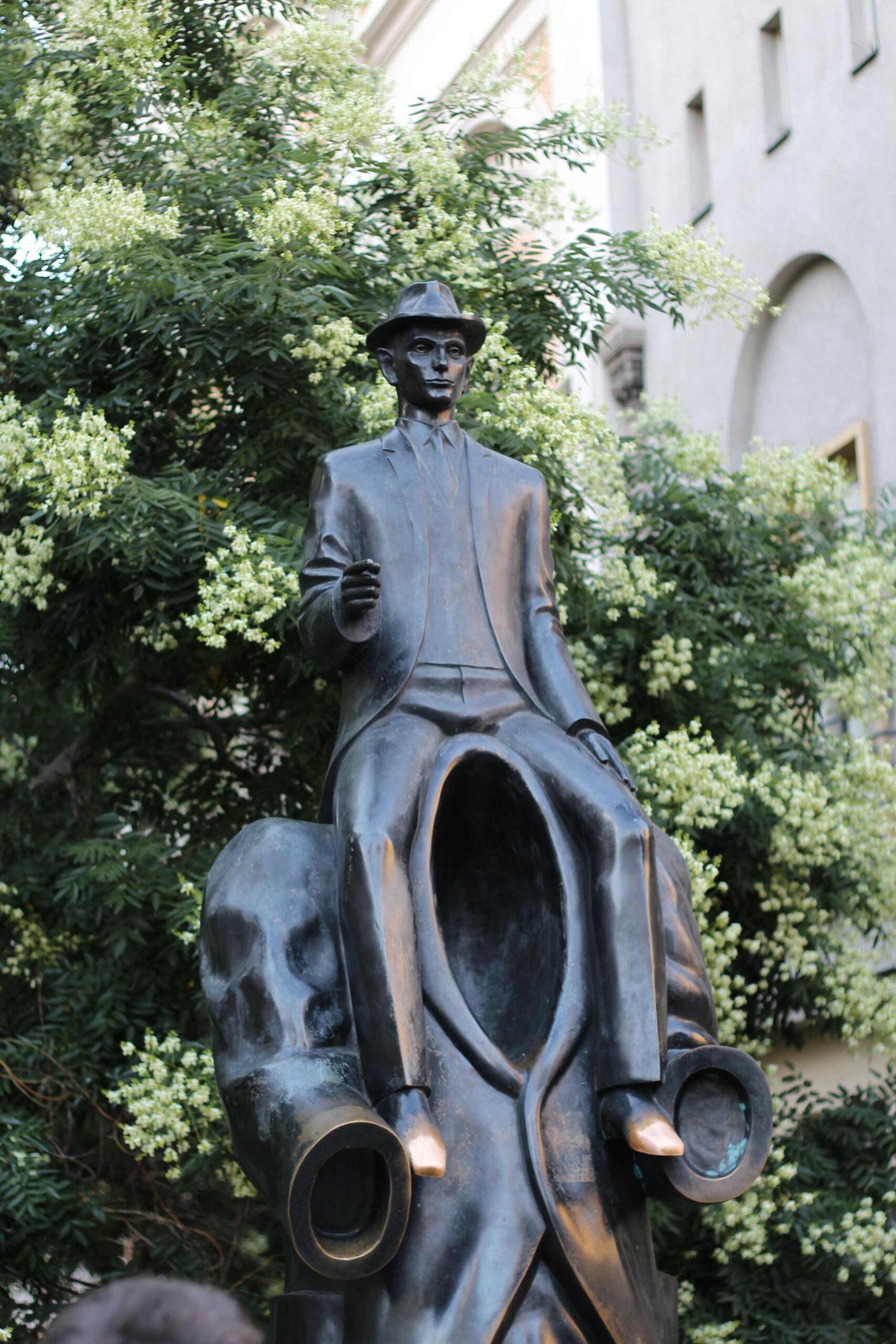 Ein großer blühender Strauch, davor eine Bronzestatue von Franz Kafka.