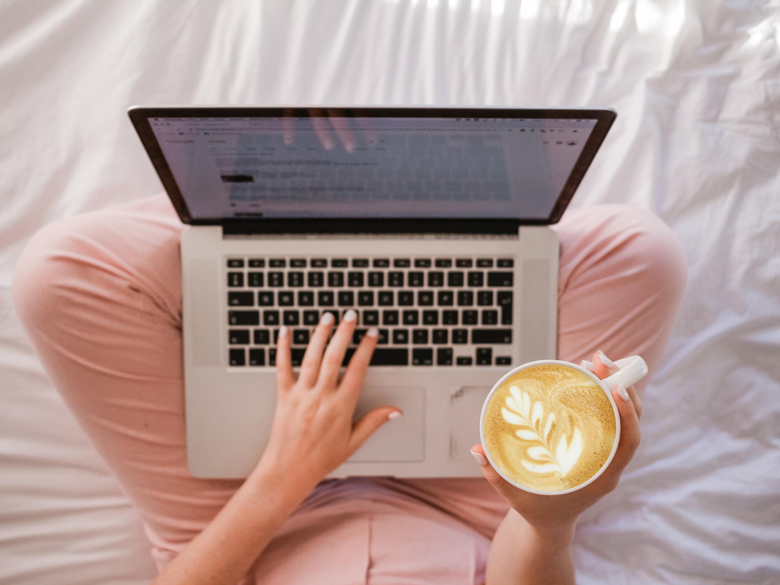 Die Beine einer Frau, die im Schneidersitz auf einem Bett sitzt. Man sieht von oben ihre Hände, sie tippt auf einem Laptop und hat einen Kaffee in der rechten Hand.