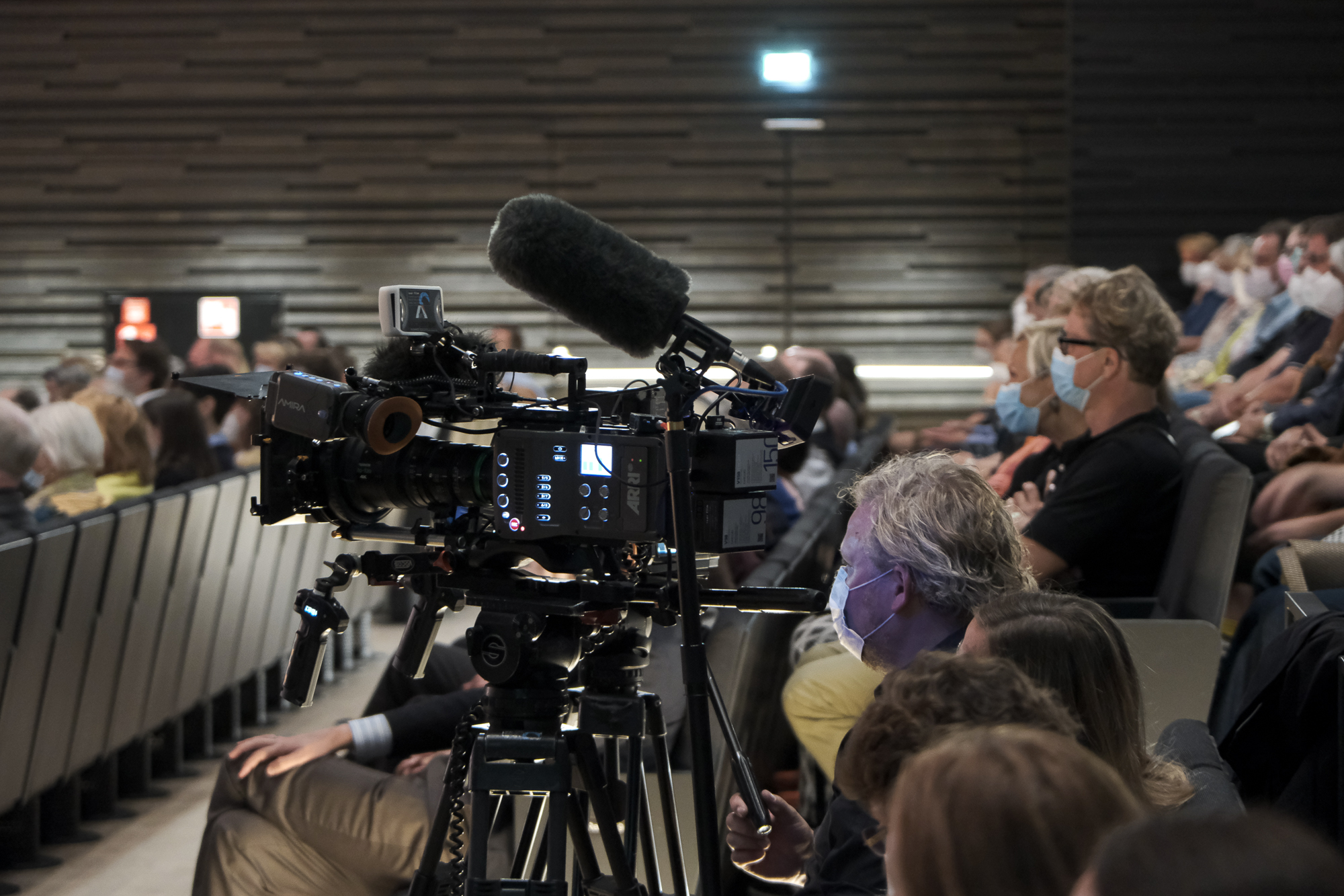 Rows of seats in the Isarphilharmonie with people, a TV camera with a microphone is set up between them
