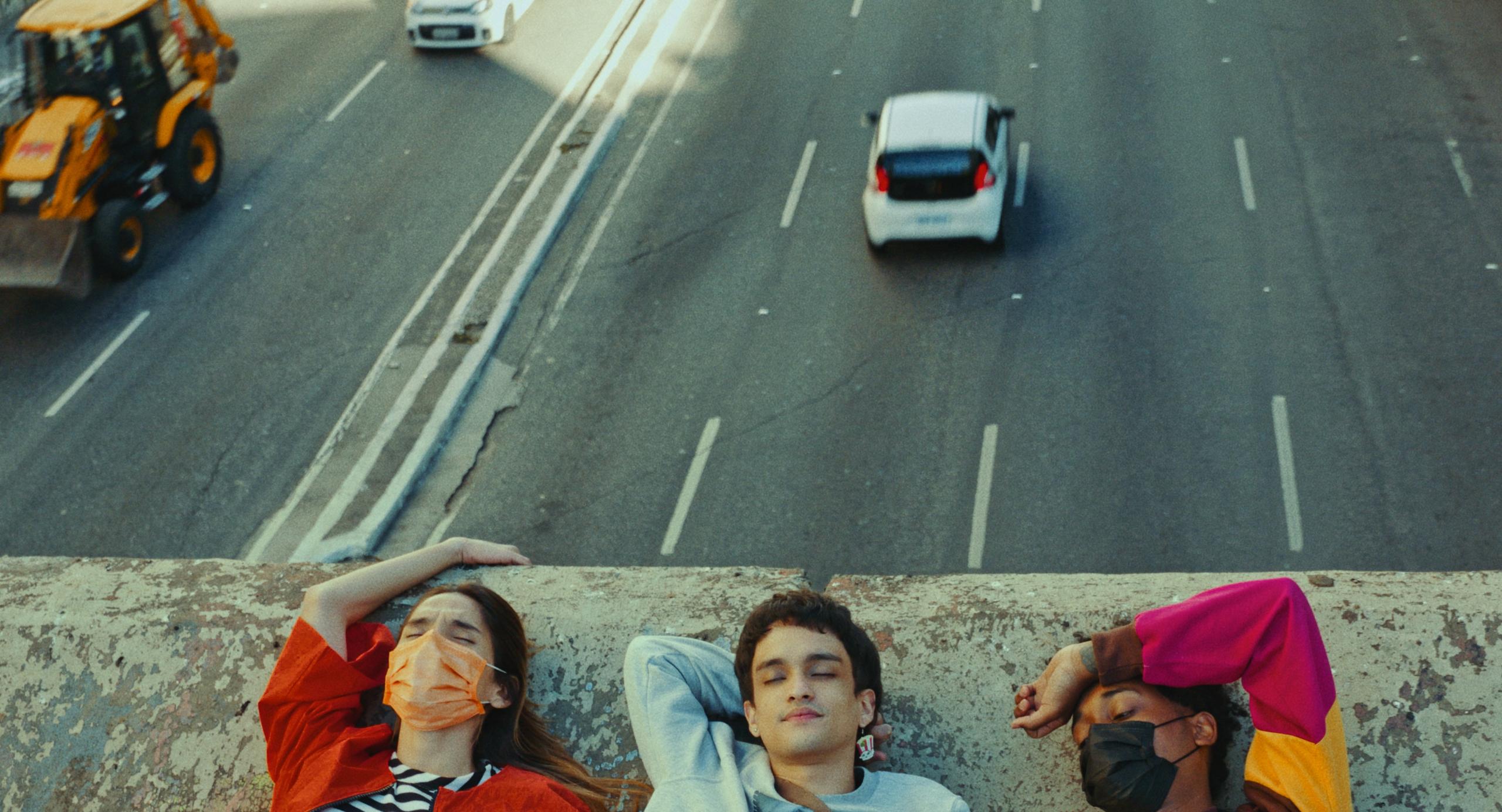 Three young people are lying on the edge of a bridge. Cars are driving below them on a multi-lane road.